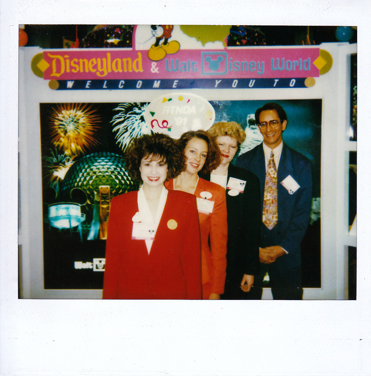 4 Disney Employees stand in a line at the Disneyland / Walt Disney World booth at the Radio-Television News Director's Association annual conference. Jennifer in a red suit, Barbara in an orange suit, Johanna in a green suit, and Glenn in a blue suit